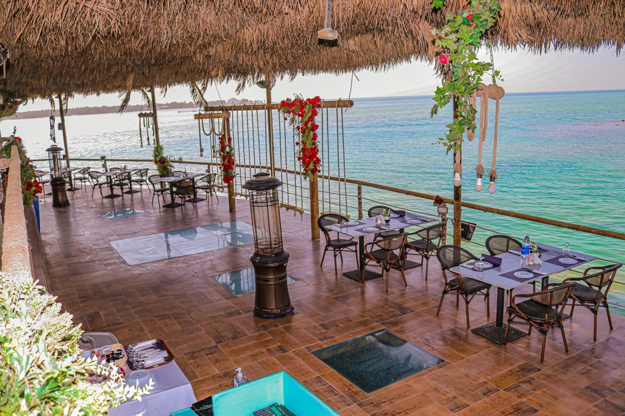 מנאמה Lagoona Beach Luxury Resort And Spa מראה חיצוני תמונה The photo depicts a coastal restaurant or dining area with an open-air design. It features a wooden floor and tables set for dining, surrounded by chairs. The space is partially covered by a thatched roof, providing shade while allowing for a view of