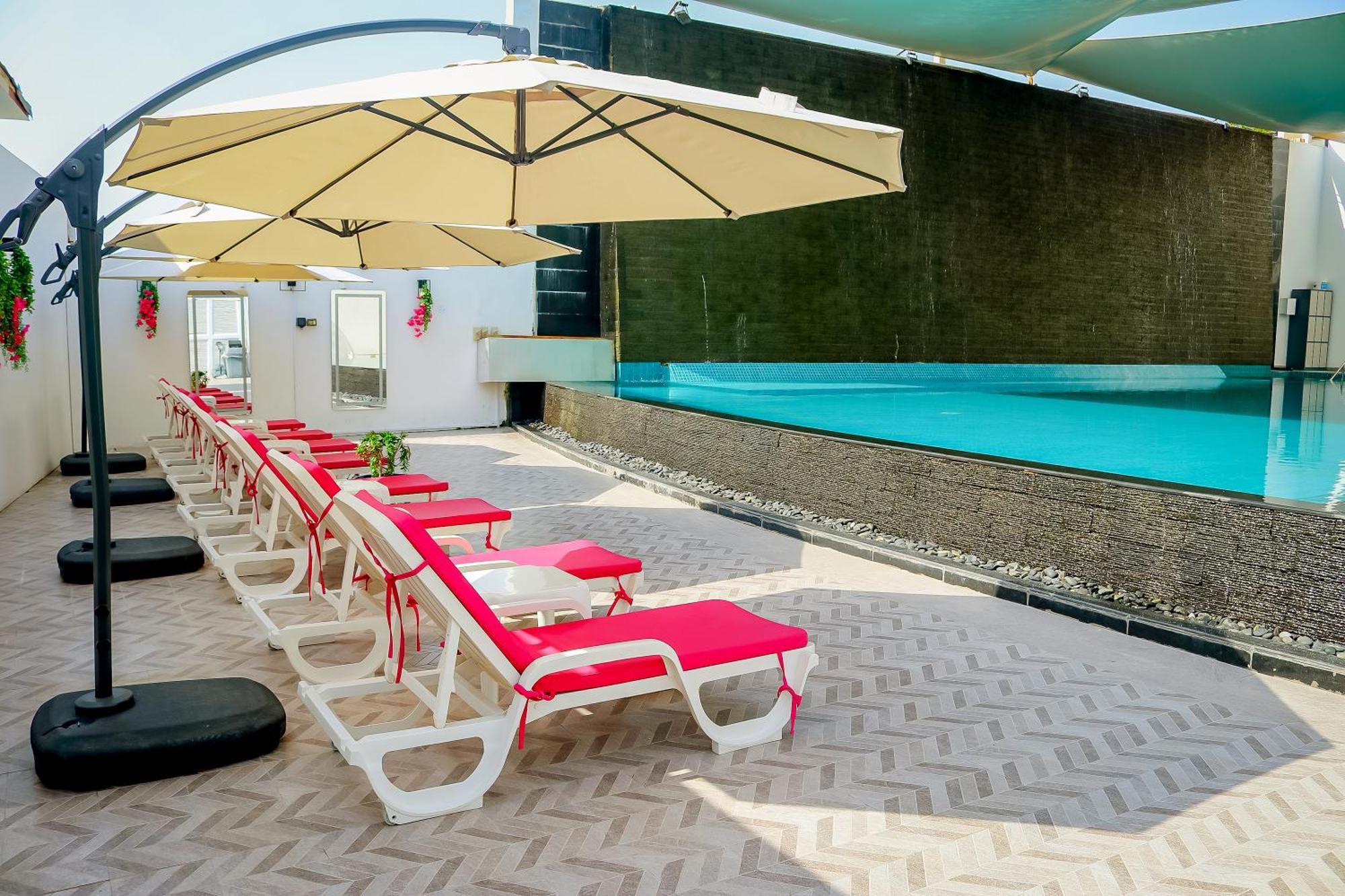 מנאמה Lagoona Beach Luxury Resort And Spa מראה חיצוני תמונה The photo shows a sunny poolside area with several lounge chairs arranged in a row. Each chair features red cushions and is positioned under large, beige umbrellas for shade. In the background, there is a beautiful swimming pool with clear blue water