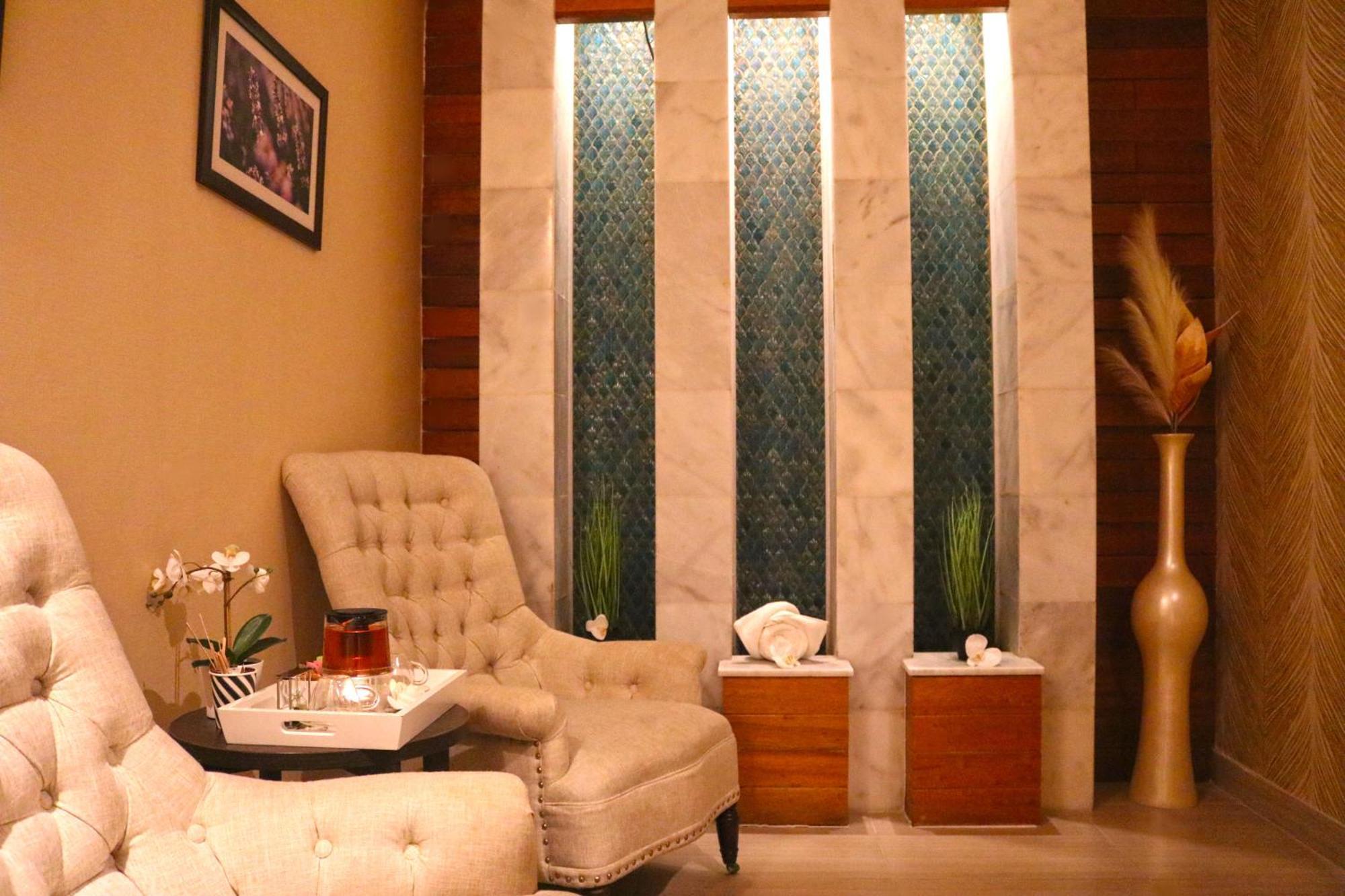מנאמה Lagoona Beach Luxury Resort And Spa מראה חיצוני תמונה The image shows a relaxing interior space, likely a waiting area or lounge. There are two plush, beige armchairs positioned near a small table, which holds a glass pitcher and a cup. The background features tall, elegant windows with decorative blue 