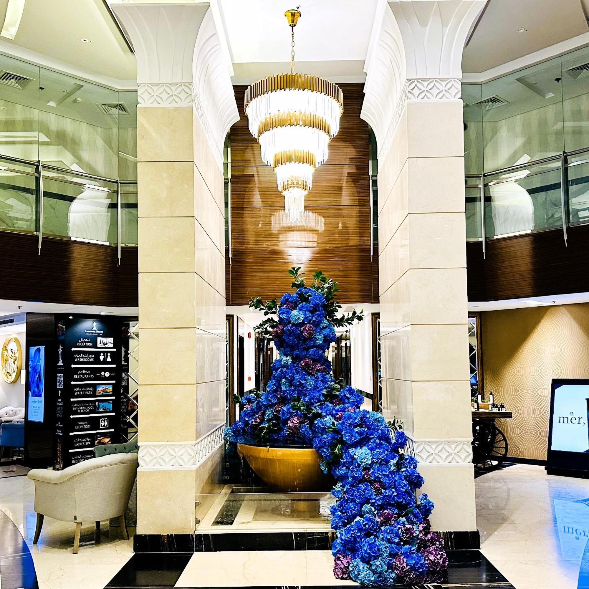מנאמה Lagoona Beach Luxury Resort And Spa מראה חיצוני תמונה The photo depicts an elegant lobby or entrance area of a building. Central to the scene is a large floral arrangement featuring vibrant blue flowers. This arrangement is positioned in a decorative bowl. Above it hangs a stylish chandelier with a laye