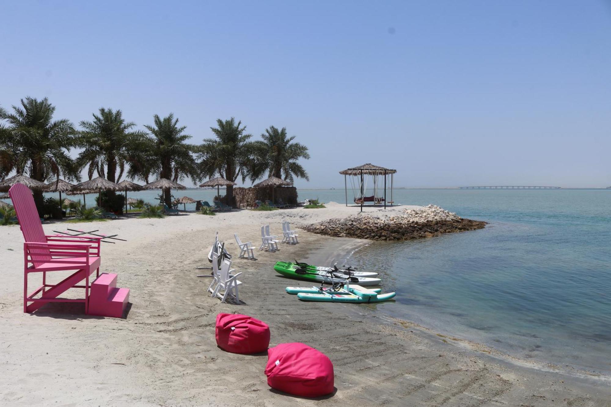 מנאמה Lagoona Beach Luxury Resort And Spa מראה חיצוני תמונה The photo depicts a serene beach scene with clear blue water and a sunny sky. In the foreground, there are colorful pink bean bags and a large pink chair on the sand. A few white chairs and loungers are positioned near the water's edge. To the side, 