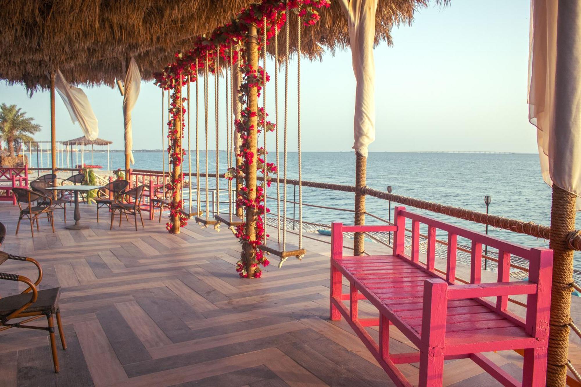 מנאמה Lagoona Beach Luxury Resort And Spa מראה חיצוני תמונה The photo depicts a scenic outdoor area by the water, with a wooden deck and a thatched roof. Two swings adorned with flowers are hanging from the ceiling, providing a charming touch to the space. In the foreground, there is a bright pink bench, addi