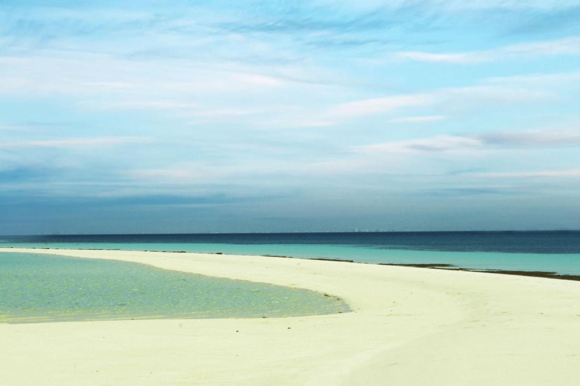 מנאמה Lagoona Beach Luxury Resort And Spa מראה חיצוני תמונה The photo depicts a serene beach scene with a gentle curve of sandy shore leading into shallow turquoise waters. The calm ocean stretches into the horizon, meeting a sky filled with soft clouds in various shades of blue. The overall atmosphere is tra
