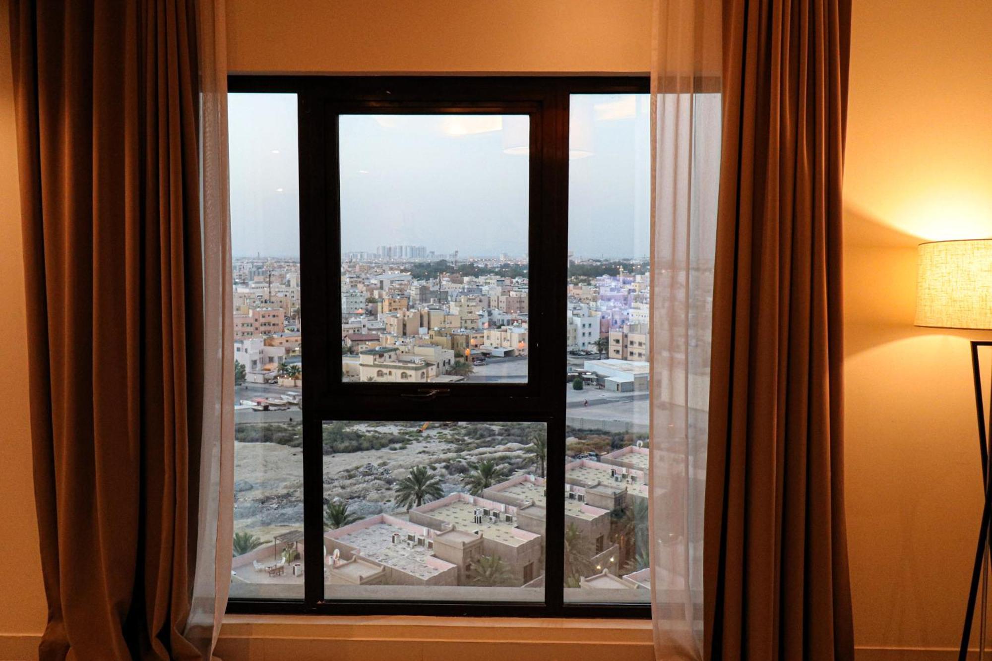 מנאמה Lagoona Beach Luxury Resort And Spa מראה חיצוני תמונה The photo shows a window with a black frame and sheer curtains. Through the window, there is a view of a cityscape, displaying a densely populated area with various buildings and rooftops. The sky appears slightly overcast, suggesting a muted lightin