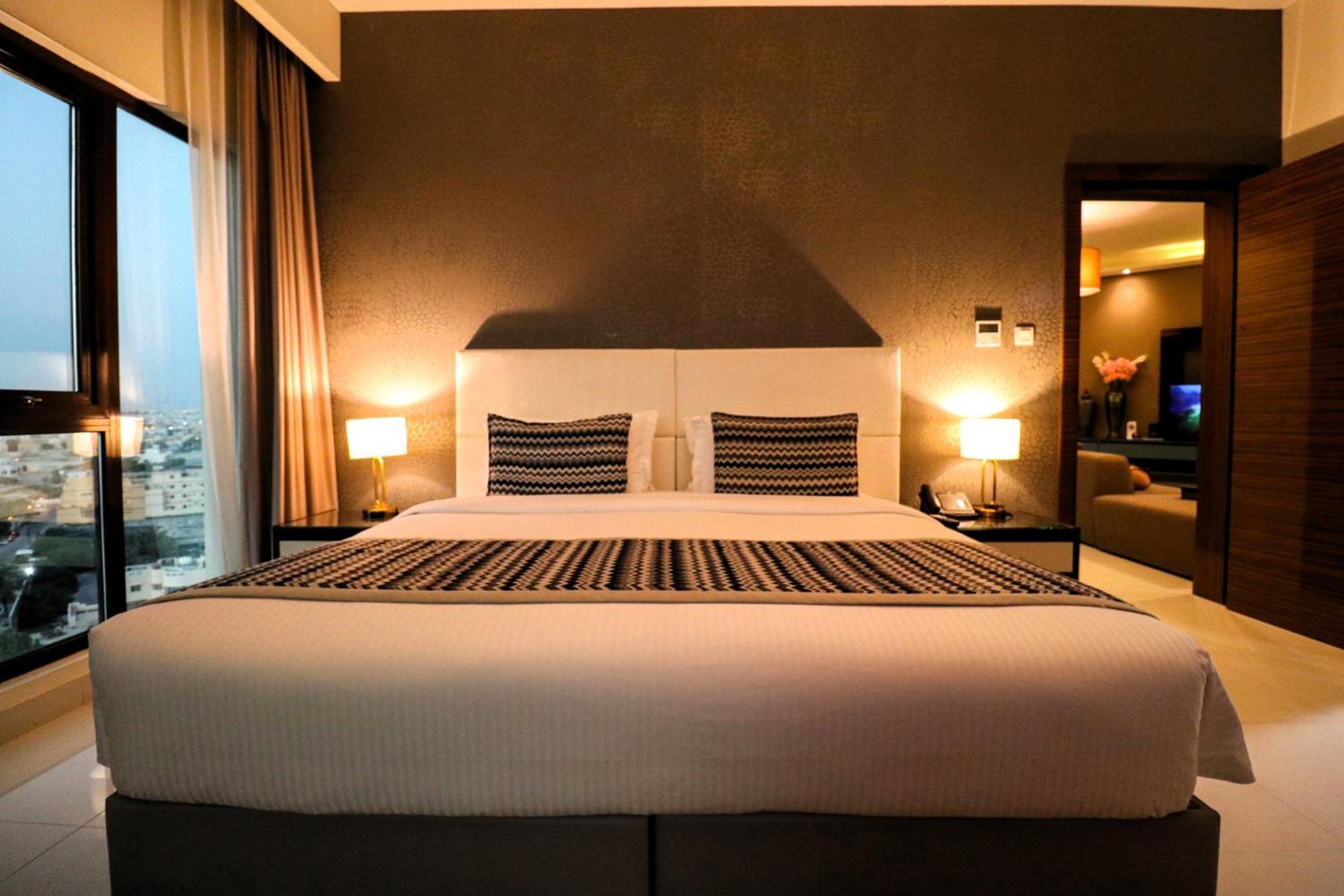 מנאמה Lagoona Beach Luxury Resort And Spa מראה חיצוני תמונה The photo depicts a modern hotel room featuring a large bed with a white comforter and two decorative pillows. The headboard is light-colored, and the walls are painted a dark hue. On either side of the bed, there are bedside tables with lamps. The r