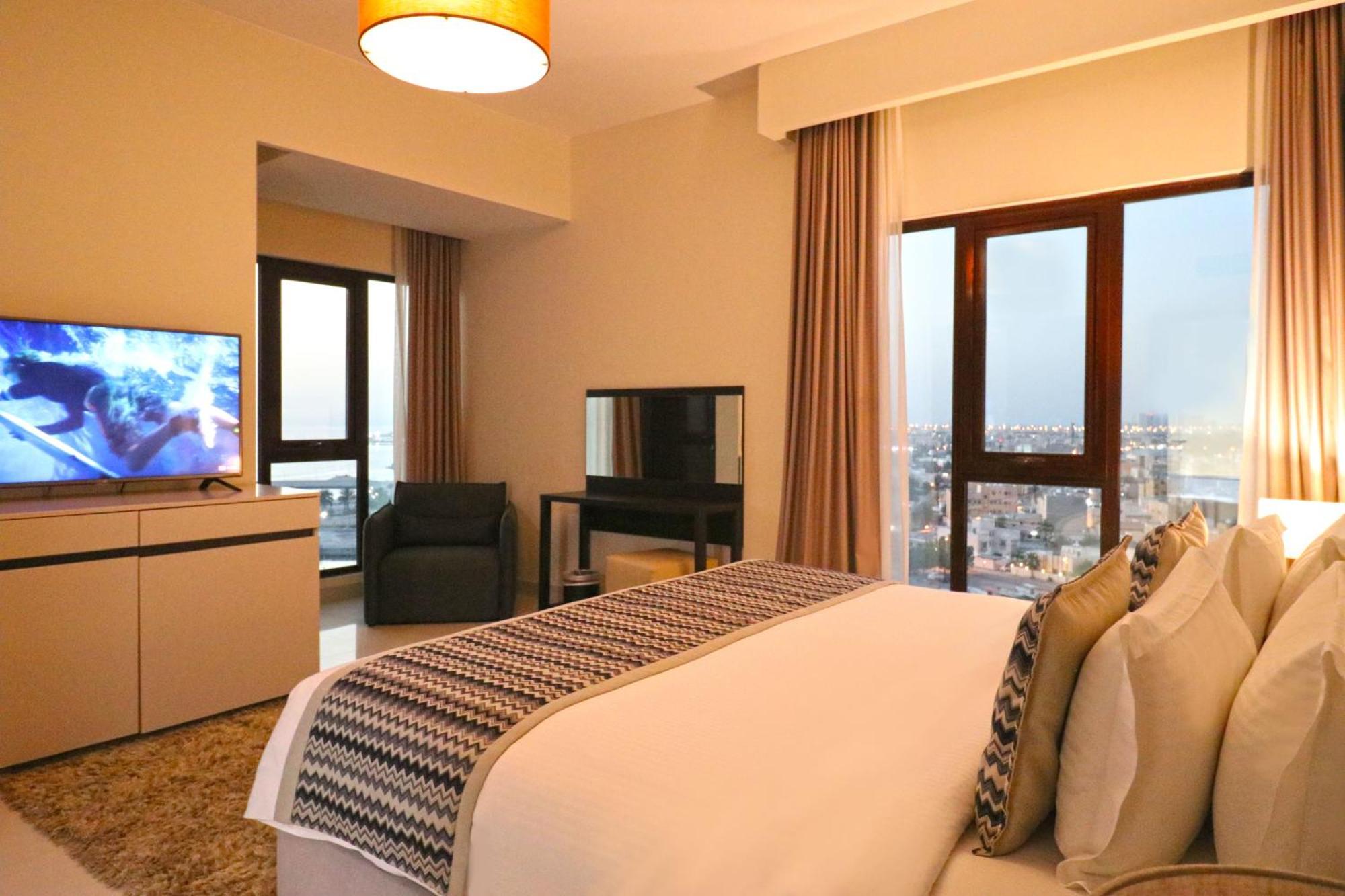 מנאמה Lagoona Beach Luxury Resort And Spa מראה חיצוני תמונה The photo shows a modern hotel room or suite. It features a neatly made bed with decorative pillows in the foreground. To the left, there is a TV on a cabinet, and further back, a comfortable chair is situated in front of a large window that likely o