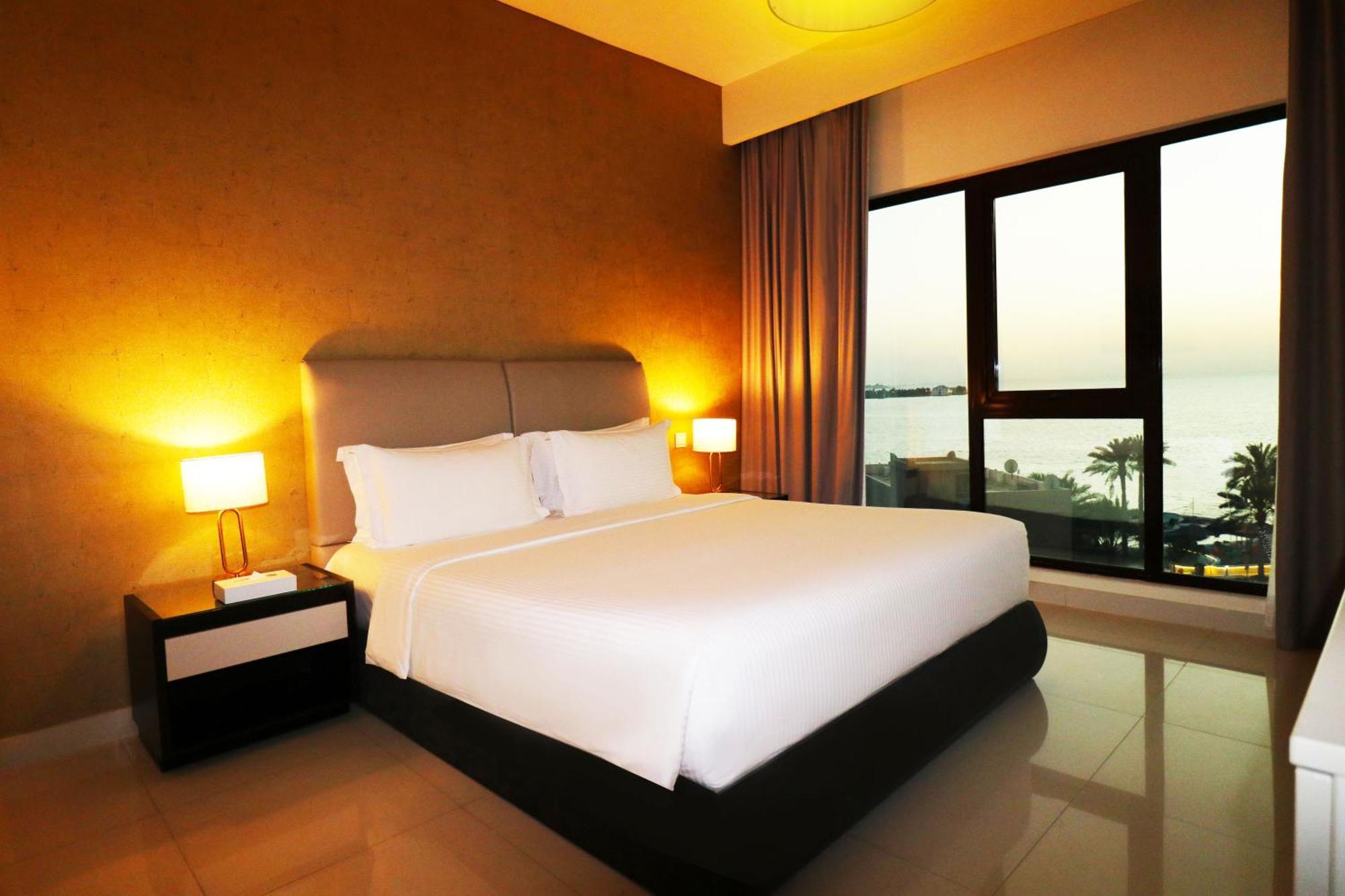 מנאמה Lagoona Beach Luxury Resort And Spa מראה חיצוני תמונה The photo shows a modern hotel bedroom with a large, comfortable bed dressed in white linens. The room features warm lighting, including bedside lamps, creating a cozy atmosphere. There are large windows with a view of the outdoors, possibly overlook