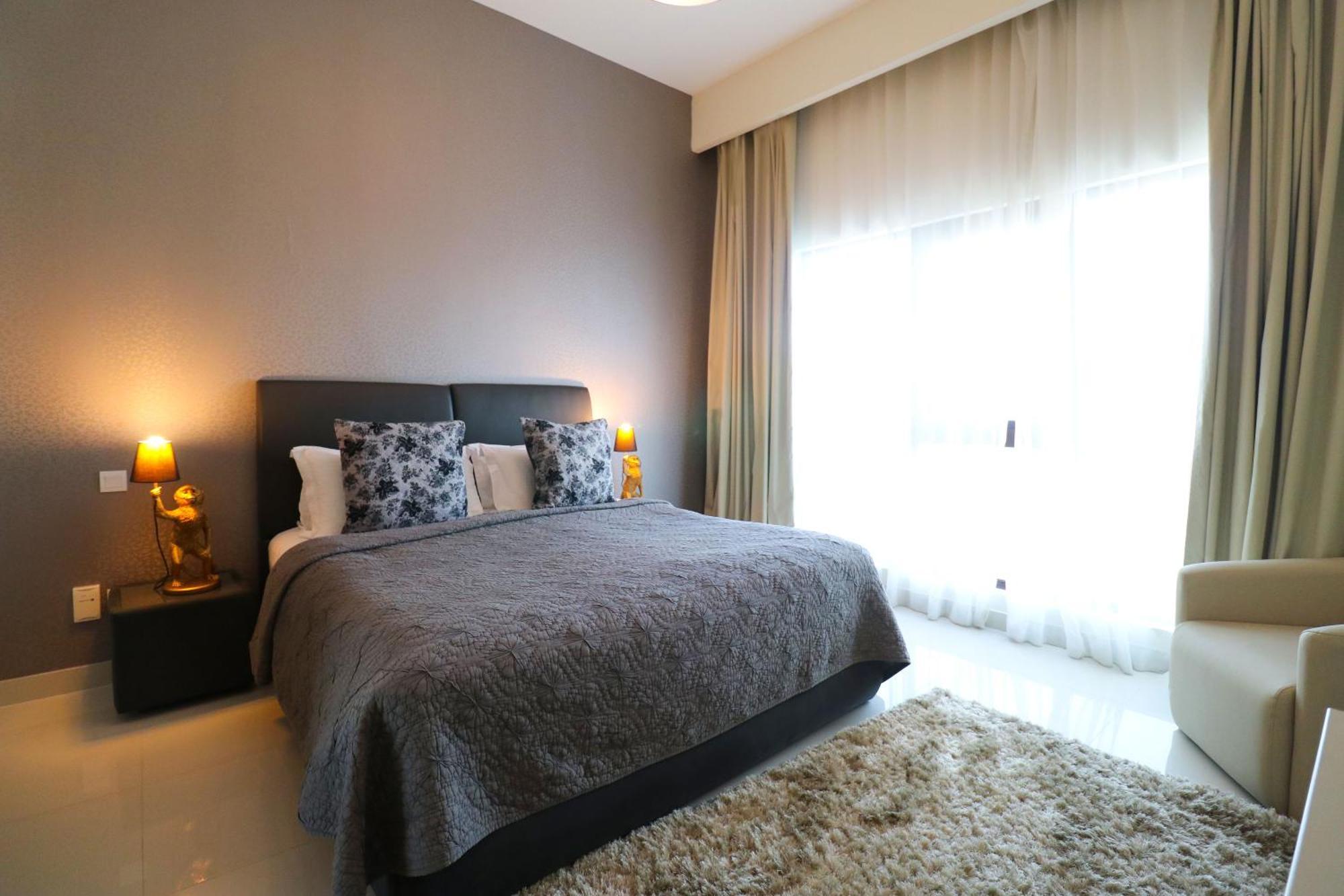 מנאמה Lagoona Beach Luxury Resort And Spa מראה חיצוני תמונה The photo shows a stylish bedroom with a modern design. It features a large bed with a dark gray bedspread and decorative pillows. On either side of the bed are two wooden nightstands, each with a lamp. There is a large window covered with light, flo